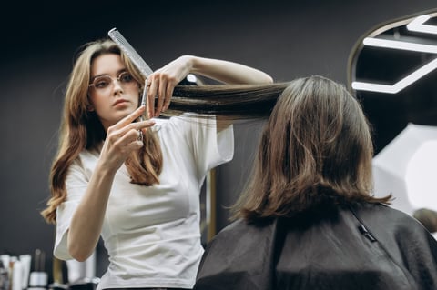 Professional female barber cutting long hair of client with scissors during hairstyling session in modern dark barbershop interior