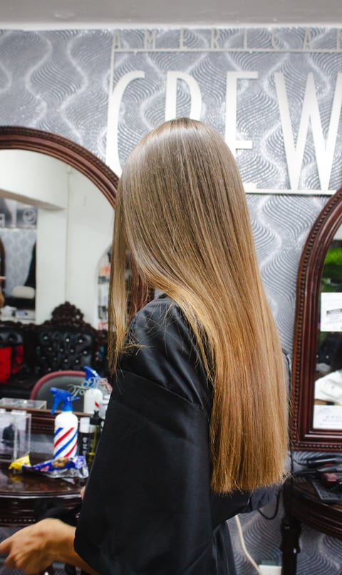 Woman with long straight brown hair in a black jacket seen from behind at a salon station with mirrors and hair styling tools