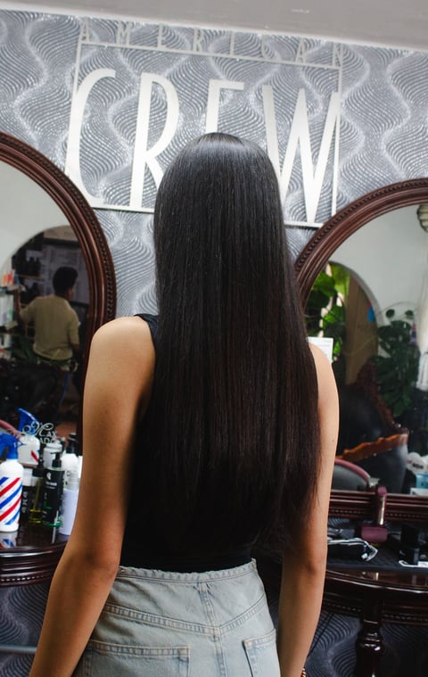 Woman with long black hair viewed from behind in a salon, facing a dual mirror with a braided salon sign above